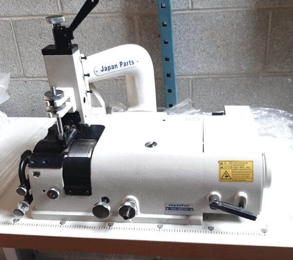Dixie Tailoring Supply The JapanFuji GT801H Heavy Skiver Sewing Machine, brand new and labeled "Japan Parts" and "JapanFuji," is set on a white table against a concrete wall, featuring visible control knobs and warning labels. Tailoring Supplies