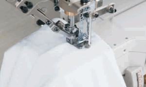 Dixie Tailoring Supply Close-up of the Jack K4 UT Automatic Coverstitch Sewing Machine stitching white fabric, highlighting the needle, threads, and mechanical components. Tailoring Supplies