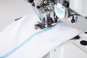 Dixie Tailoring Supply A close-up of the Jack K5 UT01GB Automatic Cylinder Arm Coverstitch Industrial Sewing Machine stitching white fabric with a blue overlock seam. Tailoring Supplies