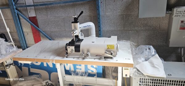 Dixie Tailoring Supply A brand new Kingmax GT801H Heavy Skiver Sewing Machine is positioned on a white work table in a warehouse, with some plastic wrapping partially removed. Tailoring Supplies