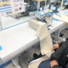 Dixie Tailoring Supply In a busy tailoring store, a person skillfully operates the JACK C4 Fully Automatic Overlock Sewing Machine, stitching a large piece of beige fabric. Tailoring Supplies