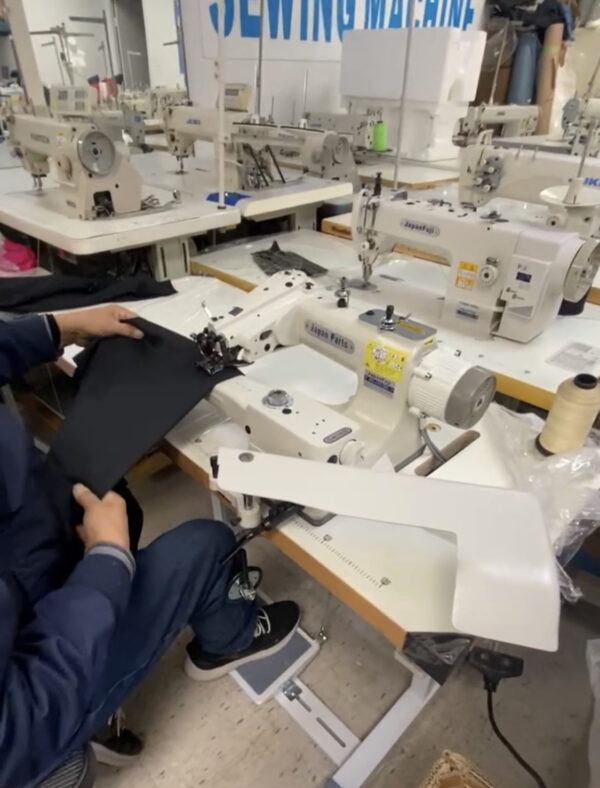 Dixie Tailoring Supply A person sews black fabric with a Japanfuji Blind Stitch Industrial Sewing Machine in a busy tailoring store, surrounded by various sewing machines, crafting exquisite tailor products. Tailoring Supplies