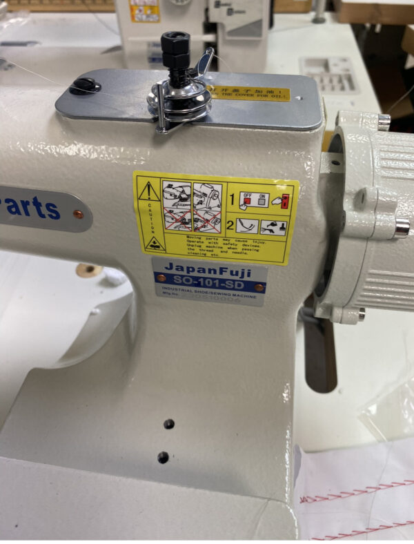 Dixie Tailoring Supply Close-up of a Japanfuji Blind Stitch Industrial Sewing Machine, ideal for any tailoring store. The image features a yellow safety instruction label and part of the stitching area in the background, perfect for showcasing quality tailor products. Tailoring Supplies