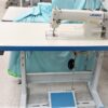 Dixie Tailoring Supply A Juki DDL 8700 brand new industrial single needle sewing machine on a stand with light blue fabric draped over it, perfectly set in a bustling tailoring store. Tailoring Supplies