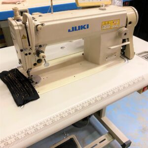 Dixie Tailoring Supply The JUKI DLN 5410N-7, a used 1-needle, needle-feed lockstitch machine, sits poised on a work table, expertly handling black fabric. Essential for any tailoring store, it ensures precise stitching and exceptional quality tailor products. Tailoring Supplies