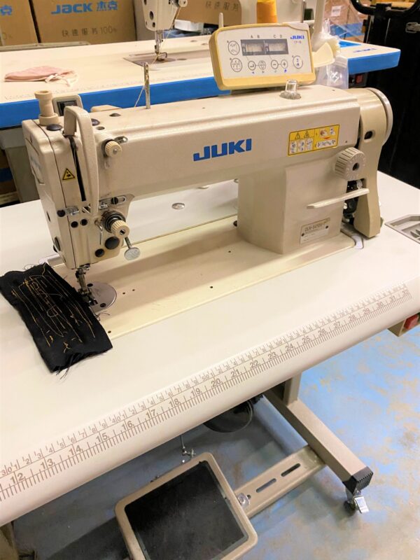 Dixie Tailoring Supply The JUKI DLN 5410N-7, a used 1-needle, needle-feed lockstitch machine, sits poised on a work table, expertly handling black fabric. Essential for any tailoring store, it ensures precise stitching and exceptional quality tailor products. Tailoring Supplies