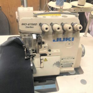 Dixie Tailoring Supply The JUKI MO 6714 Overlock Sewing Machine is expertly stitching black fabric, nestled on a table surrounded by spools of thread—an ideal addition to any tailoring store's inventory. Tailoring Supplies