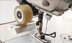 Dixie Tailoring Supply Close-up of the Jack A6F Automatic Needle Feeding Industrial Sewing Machine's needle, presser foot, and beige roller wheel attachment on the machine’s surface. Tailoring Supplies