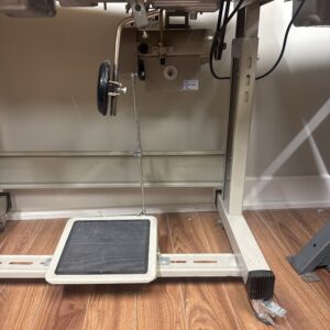 Dixie Tailoring Supply Underside of a Used Juki DDL 8700 single needle sewing machine table showing the foot pedal, wheel mechanism, power switch, and connecting rods on a wood floor. Tailoring Supplies