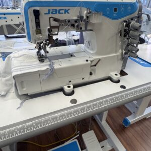 Dixie Tailoring Supply The Jack W4-D Industrial 5 Thread Flat Bed Coverstitch Sewing Machine is positioned on a work table with fabric under the needle, a foot pedal on the floor, and sunlight streaming through the windows in the background. Tailoring Supplies