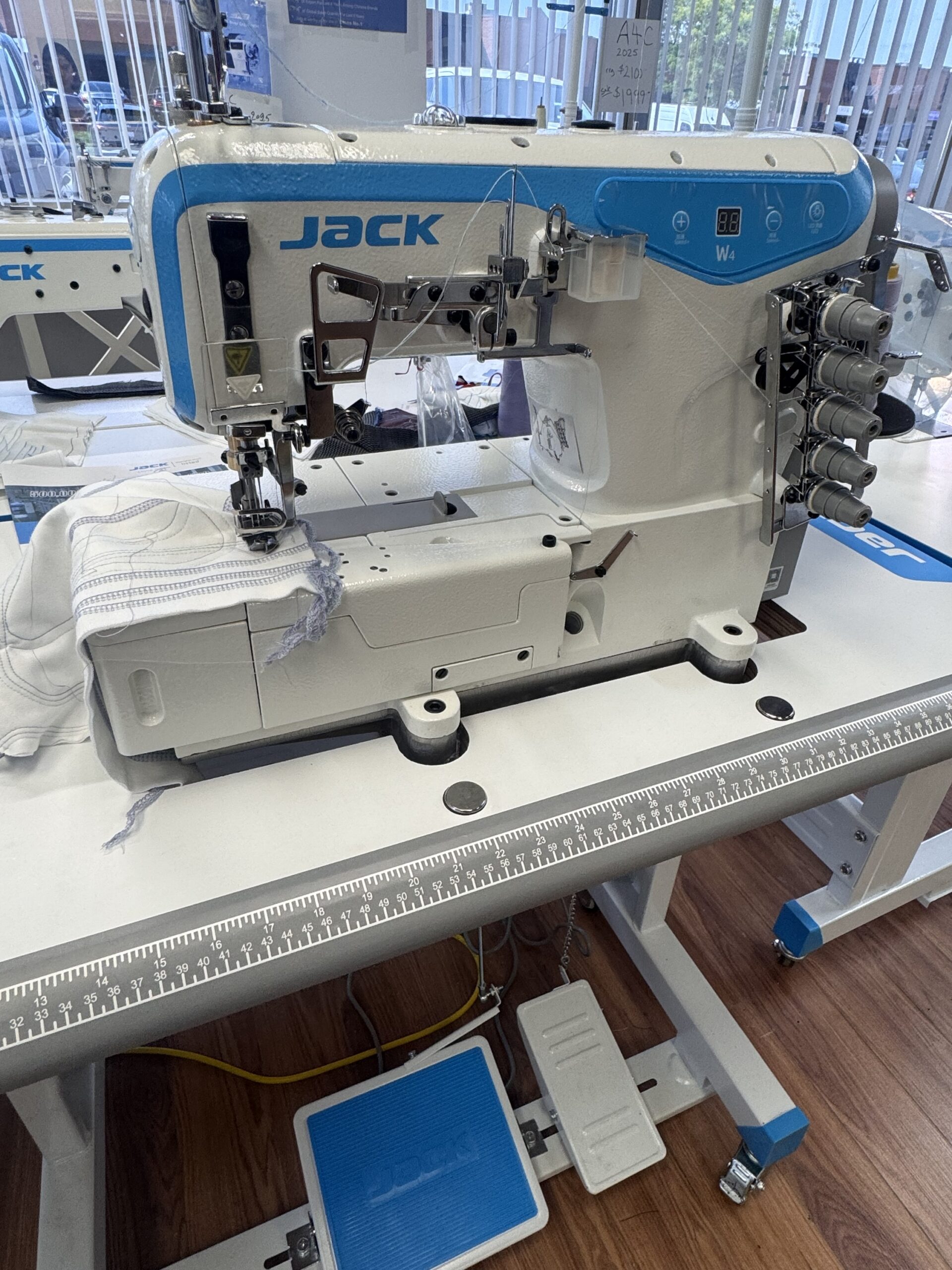 Dixie Tailoring Supply The Jack W4-D Industrial 5 Thread Flat Bed Coverstitch Sewing Machine is positioned on a work table with fabric under the needle, a foot pedal on the floor, and sunlight streaming through the windows in the background. Tailoring Supplies