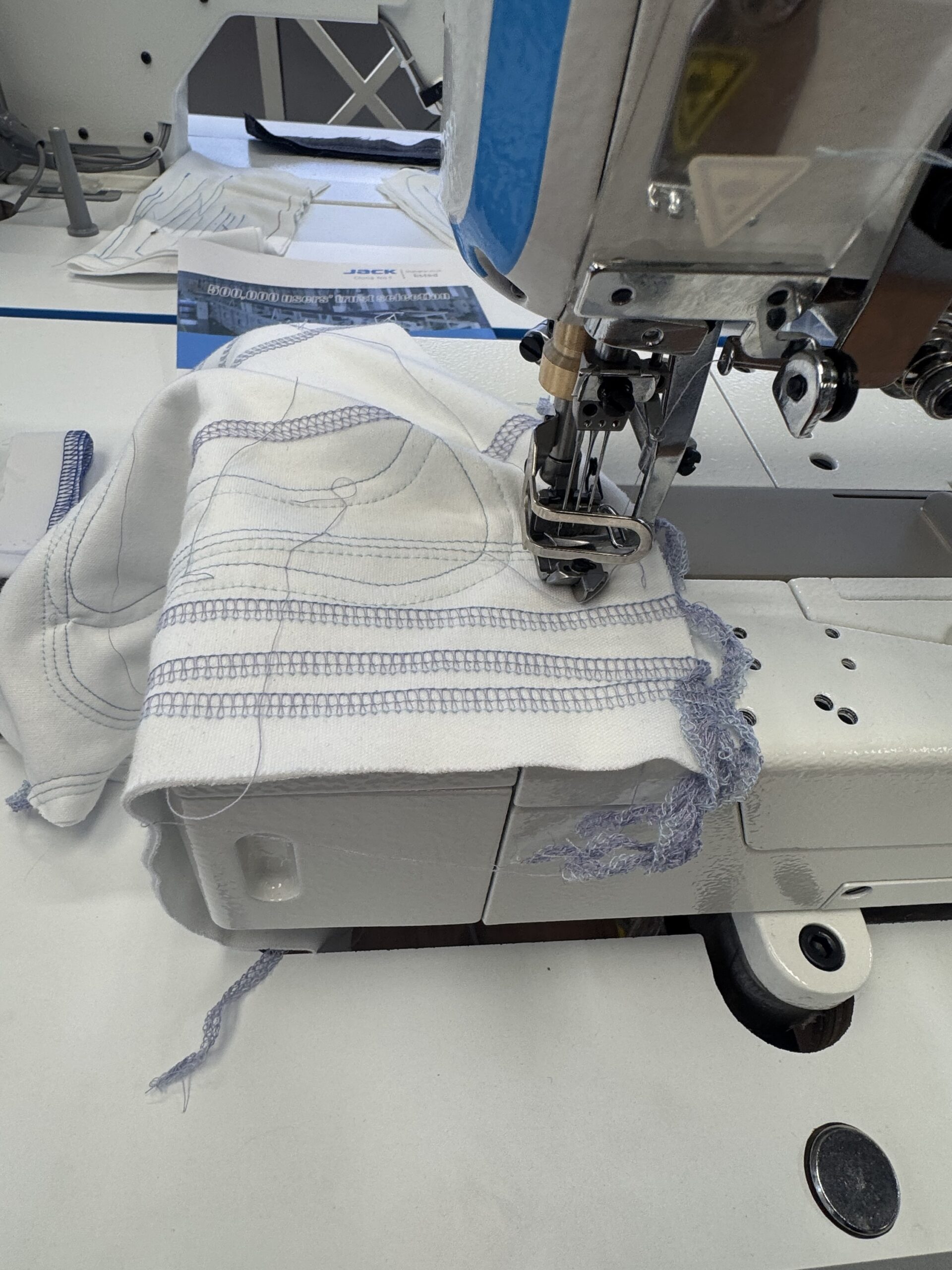 Dixie Tailoring Supply Close-up of a white fabric with blue stitching being sewn on the Jack W4-D Industrial 5 Thread Flat Bed Coverstitch Sewing Machine, with loose threads visible. Tailoring Supplies