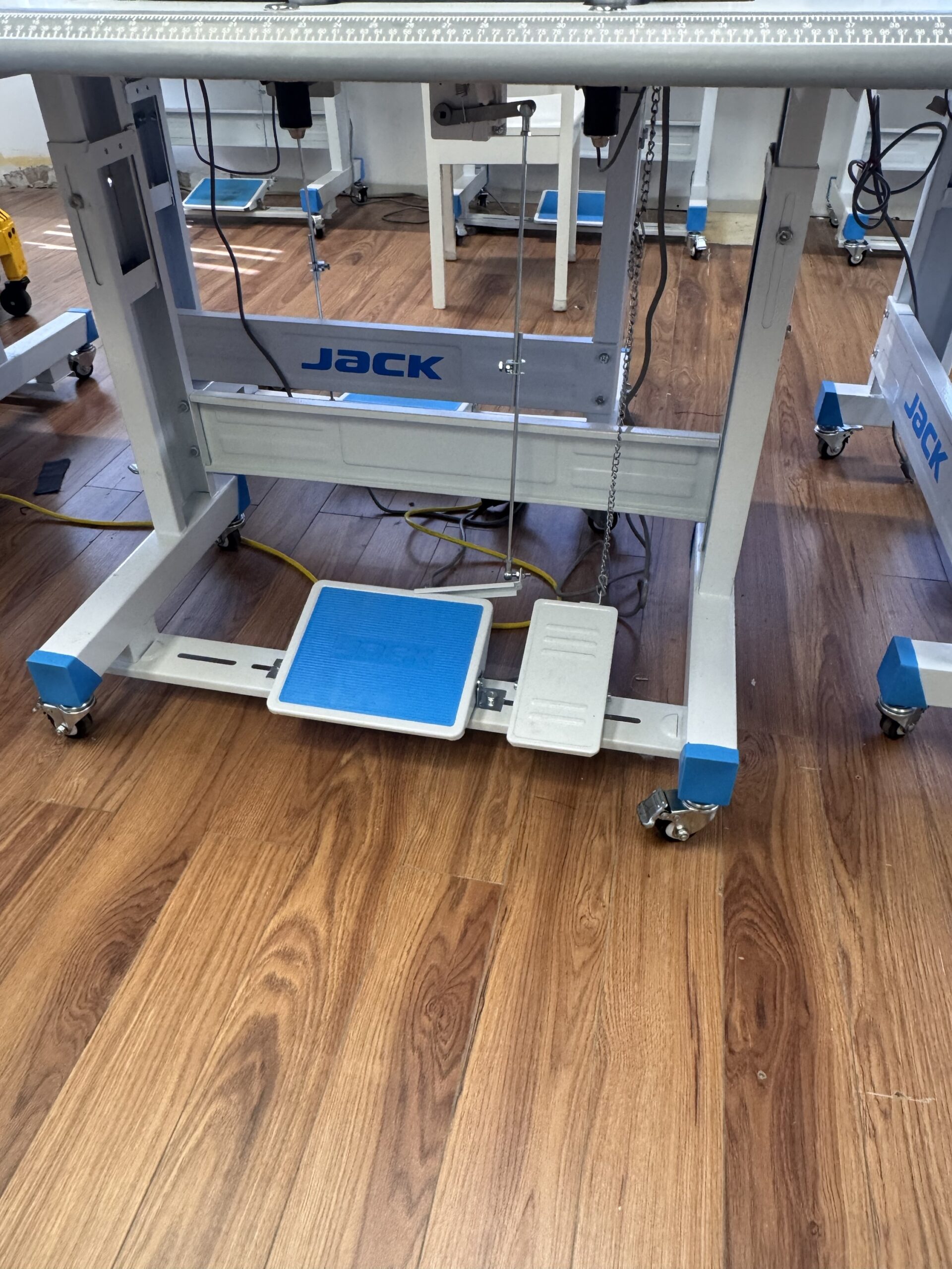 Dixie Tailoring Supply Close-up of the lower part of a Jack W4-D Industrial 5 Thread Flat Bed Coverstitch Sewing Machine table with foot pedals on a wooden floor, prominently displaying the “JACK” brand name. Tailoring Supplies