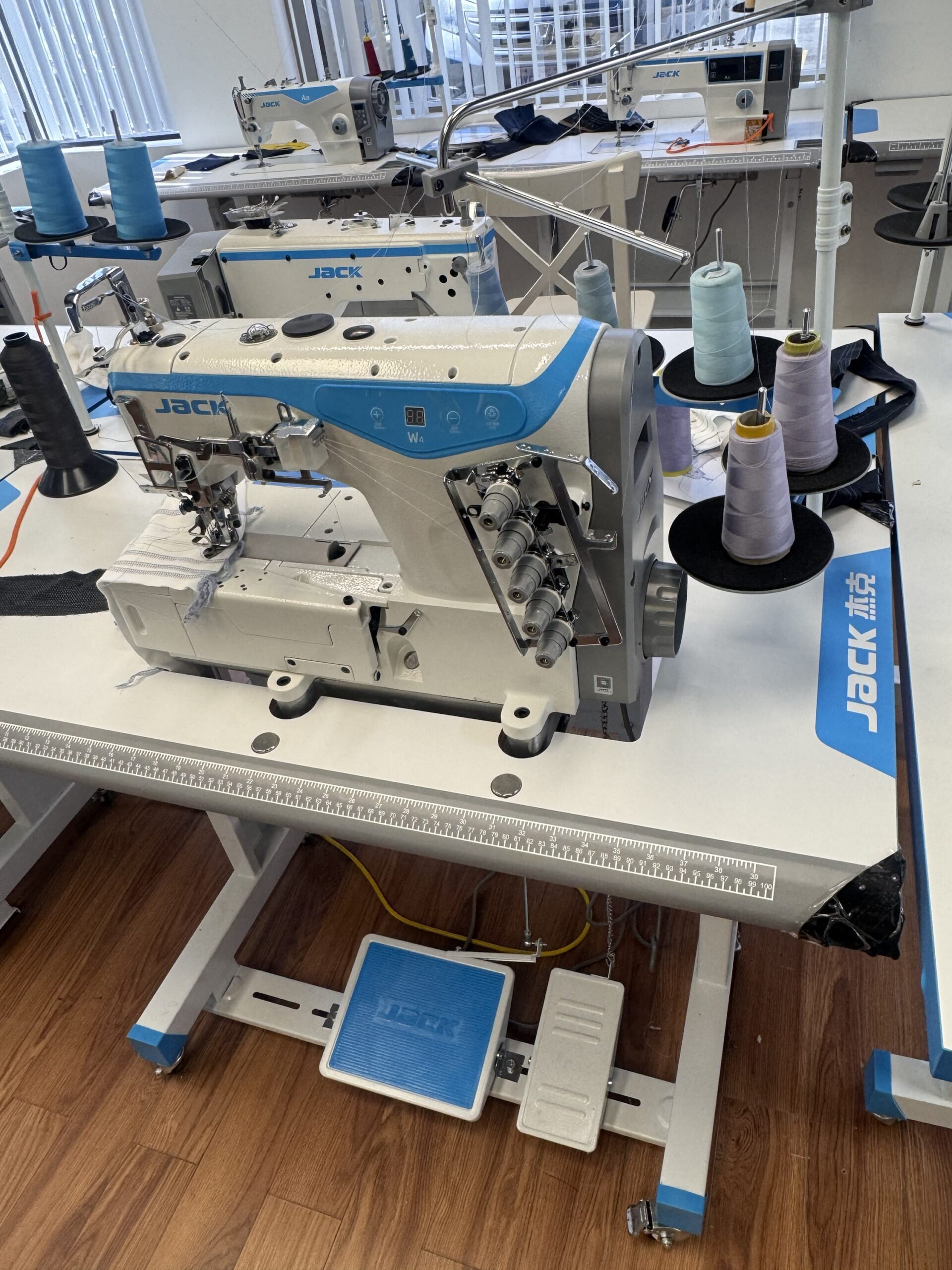 Dixie Tailoring Supply The Jack W4-D Industrial 5 Thread Flat Bed Coverstitch Sewing Machine, equipped with multiple spools of thread on a stand, is set up on a white worktable in a workspace with wooden flooring. Tailoring Supplies