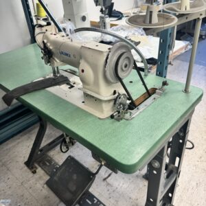 Dixie Tailoring Supply Gemsy Blind Stitch Portable Sewing Machine on a green tabletop with foot pedal, spool holders, and another sewing machine and supplies visible in the background. Tailoring Supplies