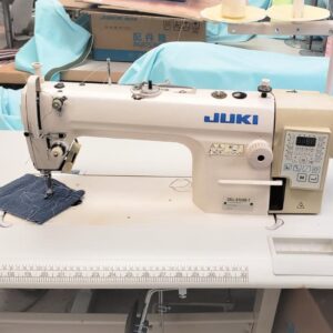 Dixie Tailoring Supply The JUKI DDL 8100-7, an industrial automatic single needle sewing machine, is set on a table with denim fabric under the needle and surrounded by tailoring supplies, creating a scene reminiscent of a bustling tailoring store. Tailoring Supplies