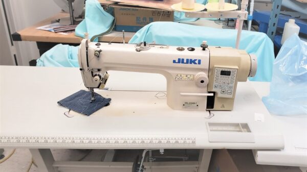 Dixie Tailoring Supply The JUKI DDL 8100-7, an industrial automatic single needle sewing machine, is set on a table with denim fabric under the needle and surrounded by tailoring supplies, creating a scene reminiscent of a bustling tailoring store. Tailoring Supplies