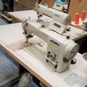 Dixie Tailoring Supply In a bustling tailoring store, two JUKI DDL 9000B sewing machines sit on a workshop table. One machine busily crafts fabric amid cardboard boxes and spools of orange thread, adding to the organized chaos. Tailoring Supplies