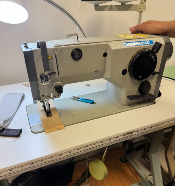 Dixie Tailoring Supply An industrial sewing machine with fabric under the needle, a pair of blue snips beside it, and a person's hand resting on the machine. Tailoring Supplies