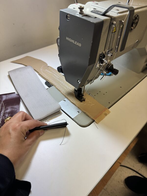 Dixie Tailoring Supply A person guiding fabric through a Highlead industrial sewing machine, with a hand holding thread near stitched material on the table. Tailoring Supplies